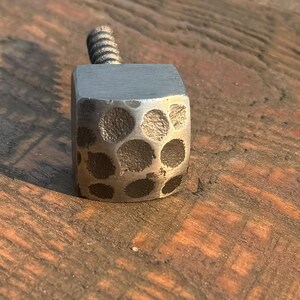 May include: A silver metal square-shaped knob with a hammered texture and a screw thread. The knob is on a wooden surface.