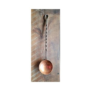May include: A copper ladle with a twisted handle. The handle is made of metal and is a light silver color. The ladle is sitting on a wooden surface.