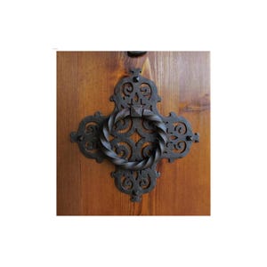 May include: A black metal door knocker with a twisted ring handle and intricate scrollwork design. The knocker is mounted on a wooden door.