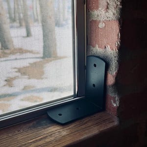 May include: Black metal corner brace mounted on a wooden window sill. The brace is attached to a brick wall.