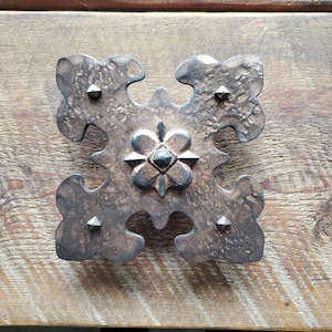May include: A dark brown metal decorative cross with a floral design in the center. The cross has a hammered texture and is accented with small metal studs.