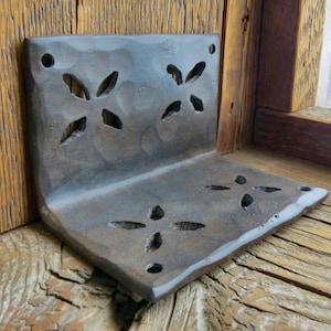 May include: A rustic, hand-forged iron shelf bracket with a decorative cutout design. The shelf bracket is made of dark metal and has a rough, textured surface.