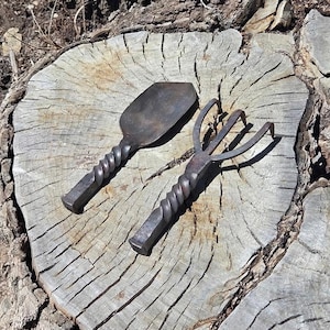May include: A dark gray metal hand trowel and a three-pronged hand cultivator. Both gardening tools feature a distinct twisted handle design and a square-shaped end, crafted from forged metal with a weathered, dark, and textured finish.