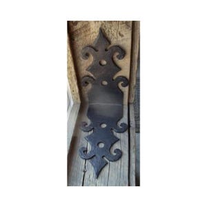 May include: Black metal decorative hinge with a scroll design. The hinge is attached to a wooden surface.