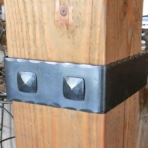 May include: A close-up of a wooden post with a black metal bracket. The bracket has two square-shaped metal accents.