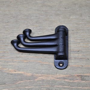 May include: Black metal wall-mounted coat rack with three hooks. The hooks are curved and have a rounded end. The rack is mounted on a wooden surface.