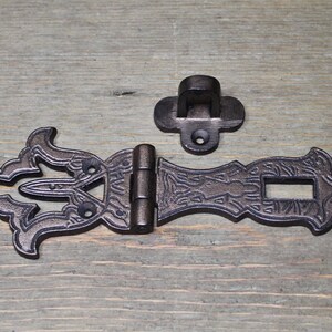 May include: A black cast iron latch with an ornate design. The latch has a hinged section and a separate hook. The latch is suitable for use on doors, cabinets, or other furniture.