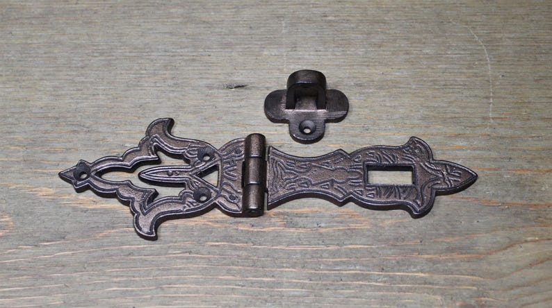 May include: A black cast iron latch with an ornate design. The latch has a hinged section and a separate hook. The latch is suitable for use on doors, cabinets, or other furniture.