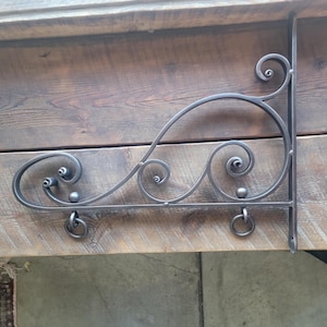 May include: A black metal sign bracket with a decorative scroll design. The bracket is mounted to a wooden wall.