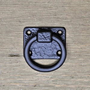May include: A black, cast iron ring pull with a square backplate. The ring is attached to the plate with a textured, square base. The backplate has four screw holes. The hardware is set against a wooden surface.