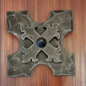 May include: A decorative metal cross with a stylized design and a raised center point. The cross is a dark gray color and has a distressed finish.