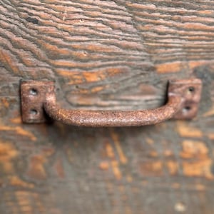 May include: A rusty metal drawer pull with a rounded handle. The pull is attached to a wooden surface with a dark brown wood grain.