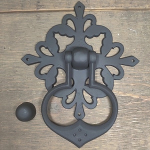 May include: Black metal door knocker with an ornate design. The knocker has a round ring and a decorative plate with a floral pattern.