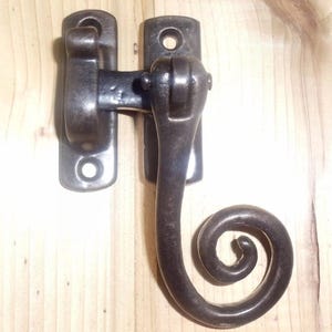 Könnte beinhalten: Ein schwarzer Fensterriegel aus Metall mit einem dekorativen Schnörkeldesign. Der Riegel ist auf einer Holzoberfläche montiert.