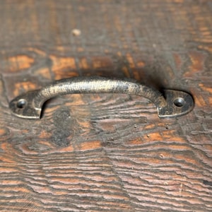 May include: A rustic, antique-style, black metal drawer pull with a slightly curved handle. The pull has a distressed finish and is mounted on a wooden surface.