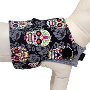May include: A dog harness cover featuring a black fabric with a repeating pattern of colorful sugar skulls and gray floral accents. The cover has a button closure and is displayed on a dog mannequin. The text "Harness Sold Separately" is visible.