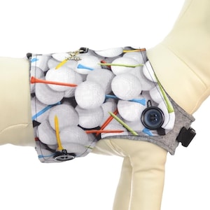 Puede incluir: Una funda para arnés de perro con un diseño de pelotas de golf. La funda es gris con un estampado de pelotas de golf en blanco, verde, naranja y azul. Tiene un cierre de botón negro y una hebilla negra. El texto "Harness Sold Separately See Description for Details" es visible.