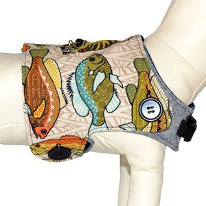 May include: A beige dog harness with a colorful fish print. The harness has a blue denim trim and a black adjustable strap. The text "Harness Sold Separately" is visible in the image.