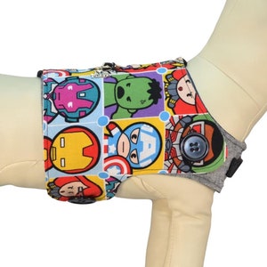 May include: A grey and blue dog vest with a colourful print of cartoon superheroes. The vest has a button closure and an adjustable strap. It's a doggy thing Pawzly.com.