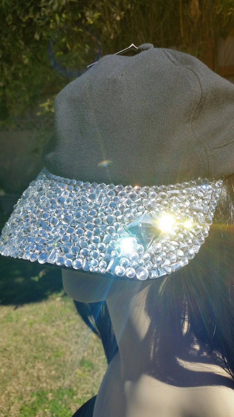 May include: A black baseball cap with a visor covered in clear rhinestones. The rhinestones are arranged in a random pattern and create a sparkling effect.