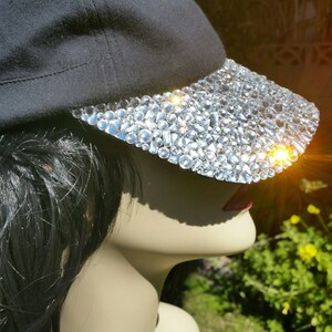 May include: A black baseball cap with a brim covered in clear rhinestones. The cap is worn on a mannequin with long black hair.