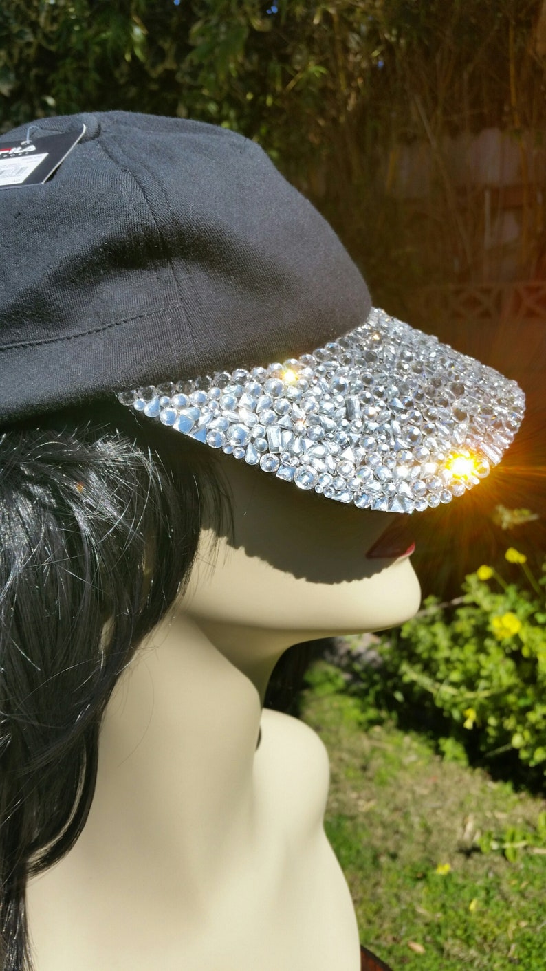 May include: A black baseball cap with a brim covered in clear rhinestones. The cap is worn on a mannequin with long black hair.