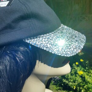 May include: A black baseball cap with a brim covered in clear rhinestones. The cap is worn on a mannequin with long black hair.