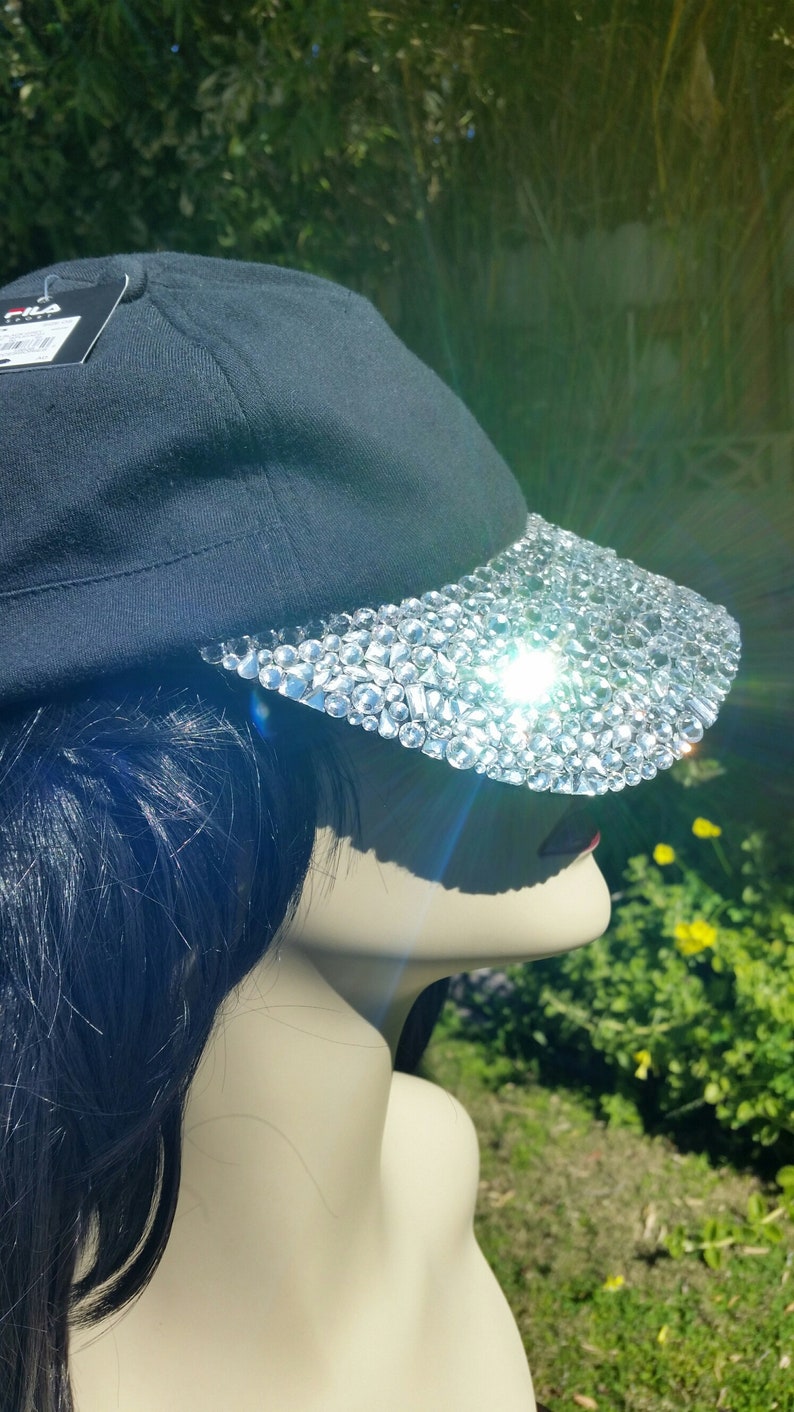 May include: A black baseball cap with a brim covered in clear rhinestones. The cap is worn on a mannequin with long black hair.