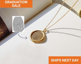 May include: A gold-colored pendant necklace with a fingerprint design. The pendant is round and has a raised edge. The necklace is on a white surface. Text in the image says "GRADUATION SALE".