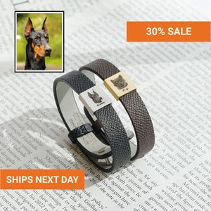 Peut inclure: Deux bracelets en cuir texturé avec des breloques métalliques carrées. Le bracelet noir possède une breloque argentée gravée d'un Doberman et du nom Kobe. Le bracelet marron a une breloque dorée gravée d'un chat et du nom Pip.