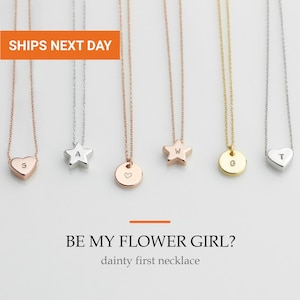 May include: Six delicate necklaces with various metal finishes and pendant shapes, including hearts, stars, and circles. Each pendant features a single stamped letter or a small heart symbol. The text BE MY FLOWER GIRL? dainty first necklace is visible.