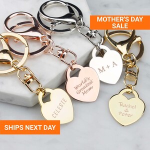 May include: Heart-shaped keychains in gold, rose gold, and silver tones. Each keychain features a heart pendant with personalized engravings, including names and the text "World's Greatest Mom." The image also includes the text "Mother's Day Sale."