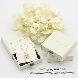 a necklace with a pendant, placed inside a white jewelry box, accompanied by a bouquet of white flowers. The box is labeled as a "handmade with love" box, and there is a note indicating that it is a " Newly upgraded, complimentary box included".