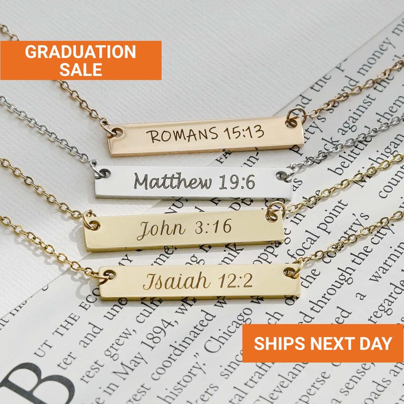 May include: Four gold and silver bar necklaces with engraved Bible verses: Romans 15:13, Matthew 19:6, John 3:16, and Isaiah 12:2. The necklaces are displayed on a printed background with the text "GRADUATION SALE".