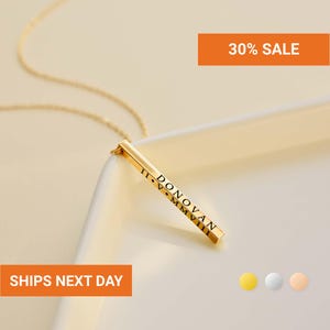 May include: A gold-toned vertical bar pendant necklace featuring the engraved name DONOVAN and the Roman numeral date II • V • MMVIII. The slender metal bar is suspended from a delicate matching chain, showcasing a polished finish and clean lines.