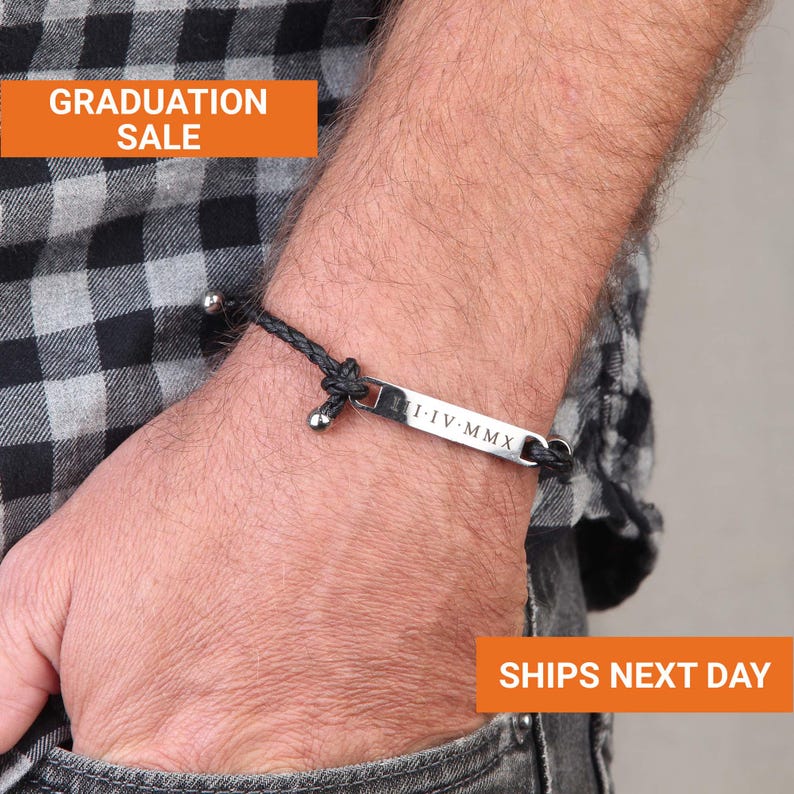 Puede incluir: Una pulsera trenzada negra con una barra plateada grabada con n&uacute;meros romanos. La pulsera se lleva en la mu&ntilde;eca. La imagen incluye el texto "GRADUATION SALE".