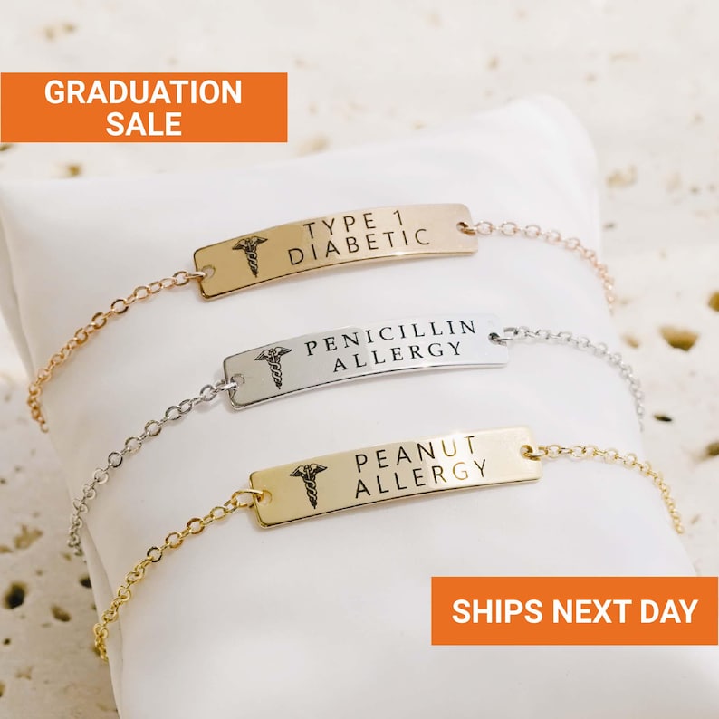Puede incluir: Tres pulseras de alerta m&eacute;dica con cadenas de oro, plata y oro rosa. Cada pulsera tiene una placa rectangular con texto grabado: "TYPE 1 DIABETIC", "PENICILLIN ALLERGY" y "PEANUT ALLERGY". Las pulseras se muestran sobre una almohada blanca.