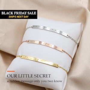 May include: Three delicate chain bracelets featuring thin, curved metal bars engraved with Morse code patterns. The set includes one silver-toned, one rose gold-toned, and one gold-toned bracelet, each displayed on a soft, off-white fabric cushion.