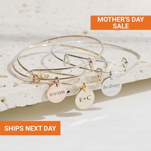 May include: Three adjustable bangles in gold, silver, and rose gold, each with a round charm. The charms are engraved with text, including "Britney", "E+C", and Roman numerals. The image includes the text "MOTHER'S DAY SALE".