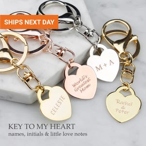 May include: Three heart-shaped metal keychains in rose gold, silver, and gold tones. Each keychain features a lobster clasp and a flat heart charm engraved with text including CELESTE, World's Greatest Mom, M + A, and Rachel & Peter.