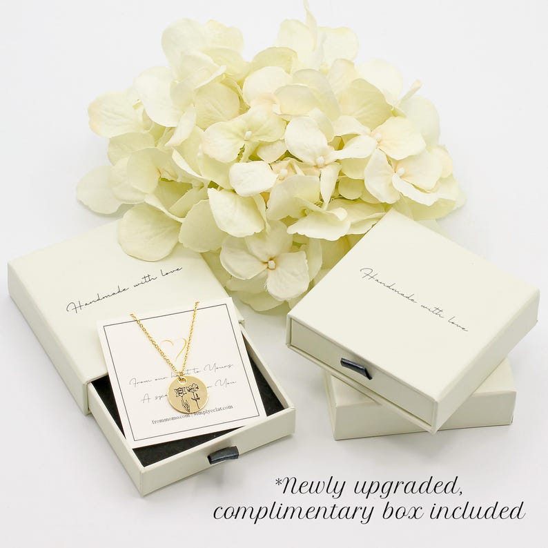 a necklace with a pendant, placed inside a white jewelry box, accompanied by a bouquet of white flowers. The box is labeled as a "handmade with love" box, and there is a note indicating that it is a " Newly upgraded, complimentary box included".