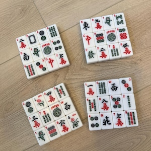 May include: Four sets of white Mahjong tiles with red and black symbols and characters. Each set is arranged in a square, displaying various numbers, suits, and special tiles like Jokers and Winds. The tiles are on a light brown wooden surface.