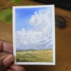 Pode incluir: Uma pintura em aquarela de uma paisagem com um céu azul com nuvens brancas fofas e pequenos pássaros pretos. A parte inferior da pintura retrata um campo de colinas onduladas amarelas e verdes.