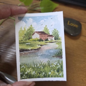 May include: A small watercolor painting of a landscape scene. The artwork depicts a house with a brown roof and white walls, surrounded by trees and a body of water. The sky is blue with a few birds. The painting is held by a person.