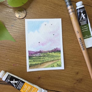 May include: A watercolor painting of a landscape with a path, green fields, and purple mountains under a blue and pink sky. The painting is on a small rectangular piece of paper. Art supplies, including paint tubes and a brush, are arranged around the painting.