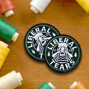 May include: Two green and black embroidered patches with white text and images. The text on the patches reads "Liberal Tears". The image on the patches is a white cartoon of a person with a paper bag over their head crying.