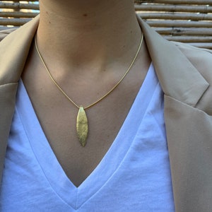 K&ouml;nnte beinhalten: Eine goldene Kettenkette mit einem goldenen Blattanh&auml;nger. Die Kette wird von einer Person mit einem wei&szlig;en Hemd und einem braunen Blazer getragen.
