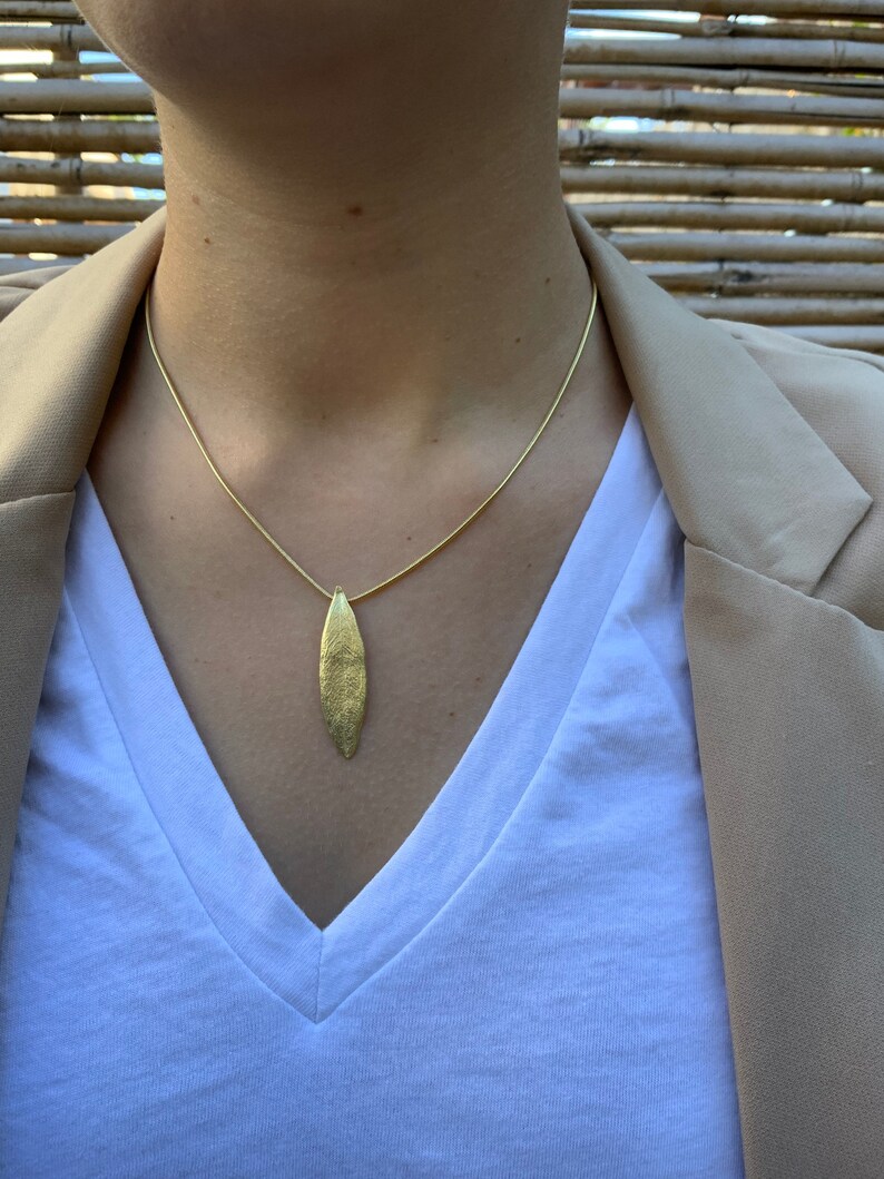 K&ouml;nnte beinhalten: Eine goldene Kettenkette mit einem goldenen Blattanh&auml;nger. Die Kette wird von einer Person mit einem wei&szlig;en Hemd und einem braunen Blazer getragen.