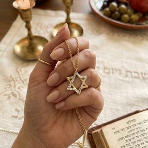 May include: A gold necklace with a Star of David pendant. The pendant has a textured silver and gold design. The necklace is held in a hand with gold rings. Lit candles and a book are in the background.