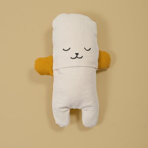 Pu&ograve; includere: Un giocattolo di peluche bianco con un viso e braccia in tessuto giallo. Il giocattolo ha un viso ricamato nero semplice con gli occhi chiusi e un piccolo naso.